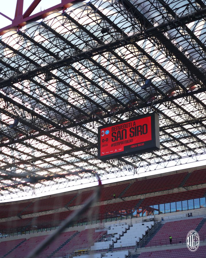 A view of the San Siro stadium with red and white seating, a large metal roof structure, and a digital scoreboard displaying "Benvenuti a San Siro" and "AC Milan" text. The field and stands are visible, with some construction elements and a logo featuring the AC Milan emblem.