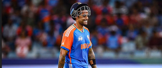 Suryakumar Yadav wearing a blue and orange cricket jersey with the BCCI logo, a helmet, and a black cap. He has tattoos on his arms and is on a cricket field with a blurred crowd in the background.