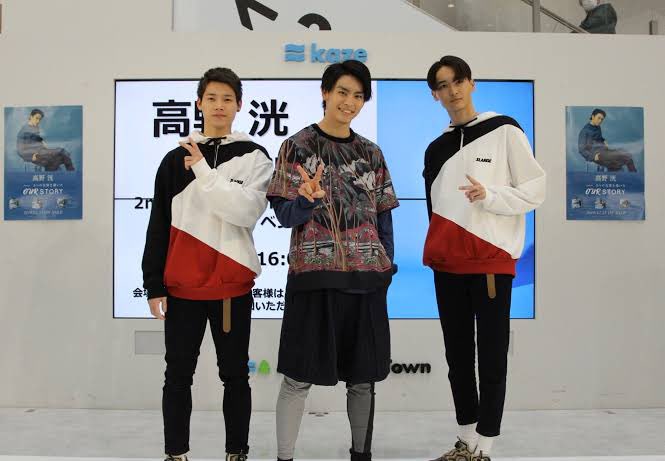 A group of people standing together on a stage in a large auditorium with a crowd in the background. They are dressed in black outfits, with one person in the center wearing a white shirt and another in a colorful jacket. A cake with decorations is placed on a table in front of them. Three individuals in matching red and white jackets stand together in front of a screen displaying text and posters.