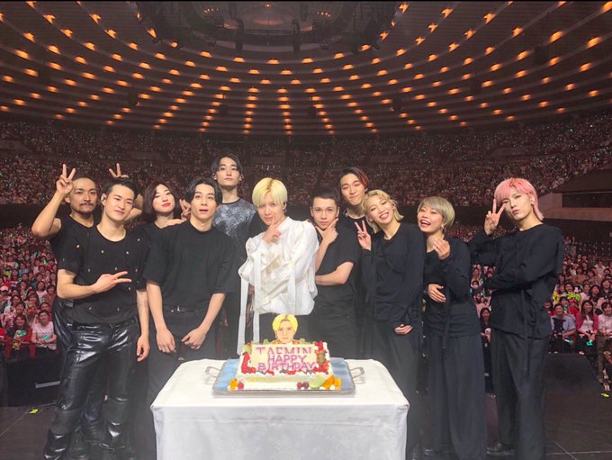 A group of people standing together on a stage in a large auditorium with a crowd in the background. They are dressed in black outfits, with one person in the center wearing a white shirt and another in a colorful jacket. A cake with decorations is placed on a table in front of them. Three individuals in matching red and white jackets stand together in front of a screen displaying text and posters.