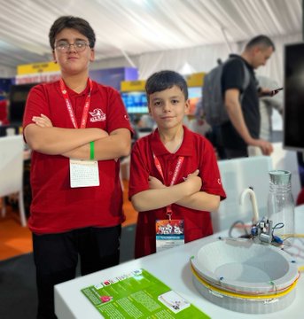 Three individuals in white shirts with arms crossed stand behind a table displaying a robot labeled "REEFSCAPE" and electronic equipment. Three individuals in red jackets with arms crossed stand behind a table displaying a robot labeled "AL-KHWARIZMI-4" and electronic devices. Two individuals in red shirts with arms crossed stand behind a table displaying a robot and a water basin. Two individuals in casual clothing with arms crossed stand behind a table displaying a screen showing "AgriAcid" and electronic equipment. The background features banners with text and colorful designs.
