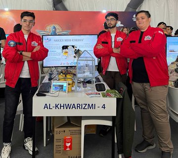 Three individuals in white shirts with arms crossed stand behind a table displaying a robot labeled "REEFSCAPE" and electronic equipment. Three individuals in red jackets with arms crossed stand behind a table displaying a robot labeled "AL-KHWARIZMI-4" and electronic devices. Two individuals in red shirts with arms crossed stand behind a table displaying a robot and a water basin. Two individuals in casual clothing with arms crossed stand behind a table displaying a screen showing "AgriAcid" and electronic equipment. The background features banners with text and colorful designs.