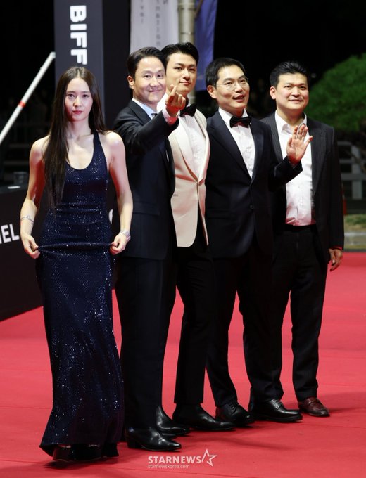 Jung Soojung in a sparkling navy blue sleeveless gown, standing on a red carpet with four men in formal attire. One man in a black tuxedo with a bow tie waves, another in a white tuxedo with a bow tie stands beside him, and two others in dark suits also wave. A "BIFF" banner and photographers are visible in the background.