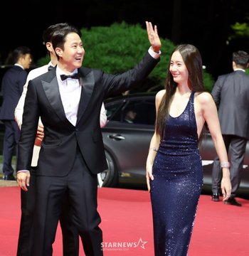 Jung Soojung in a sparkling navy blue sleeveless gown, standing on a red carpet with four men in formal attire. One man in a black tuxedo with a bow tie waves, another in a white tuxedo with a bow tie stands beside him, and two others in dark suits also wave. A "BIFF" banner and photographers are visible in the background.