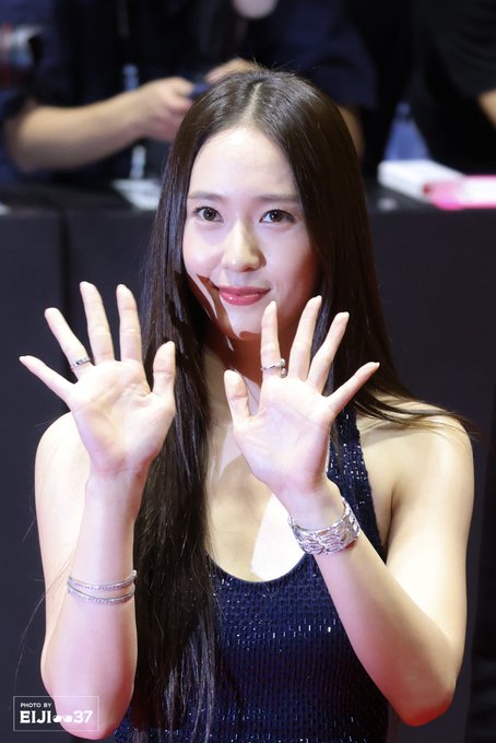 Krystal Jung standing on a red carpet, wearing a sparkling navy blue sleeveless gown with a halter neckline. Her long straight hair falls over her shoulders. She is posing with both hands raised, showing rings on her fingers and bracelets on her wrists. A watermark reading "EJI==37" is visible in the corner.