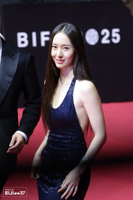 Krystal Jung standing on a red carpet, wearing a sparkling navy blue sleeveless gown with a halter neckline. Her long straight hair falls over her shoulders. She is posing with both hands raised, showing rings on her fingers and bracelets on her wrists. A watermark reading "EJI==37" is visible in the corner.