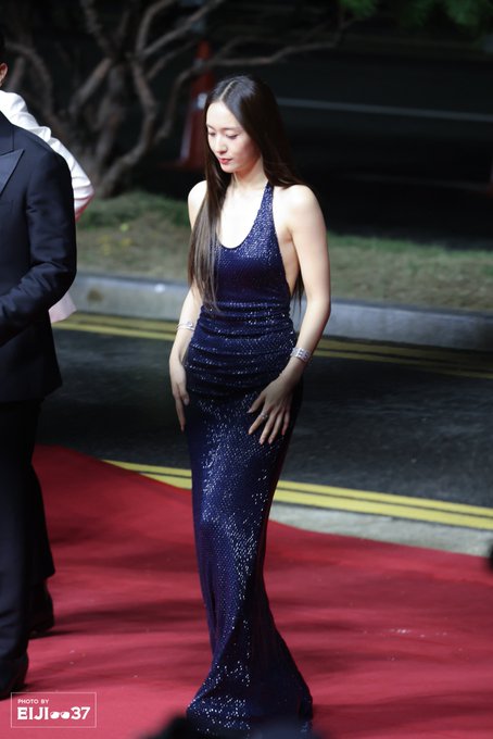 Krystal Jung wearing a sparkling navy blue halter-neck gown with a side cutout, standing on a red carpet. Her long, straight dark hair flows down her back. Several people in dark suits are visible around her, and a black backdrop with white text reading "BIFF 25" is behind her. A watermark "ELIJ_0037" is present in the lower corners of the images.