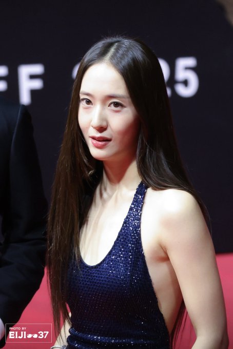 Krystal Jung wearing a sparkling navy blue halter-neck gown with a side cutout, standing on a red carpet. Her long, straight dark hair flows down her back. Several people in dark suits are visible around her, and a black backdrop with white text reading "BIFF 25" is behind her. A watermark "ELIJ_0037" is present in the lower corners of the images.