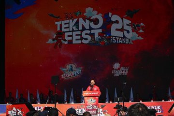 A large stage with a vibrant red backdrop displaying "TEKNOFEST 25 ISTANBUL" and logos. Pink confetti falls from above, and people in red shirts stand on stage. A crowd of attendees, some taking photos, fills the foreground. A speaker stands at a podium with a TEKNOFEST logo. Children in red jackets work on projects at a table with cardboard models and red balloons nearby.
