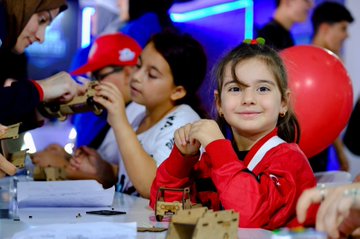 A large stage with a vibrant red backdrop displaying "TEKNOFEST 25 ISTANBUL" and logos. Pink confetti falls from above, and people in red shirts stand on stage. A crowd of attendees, some taking photos, fills the foreground. A speaker stands at a podium with a TEKNOFEST logo. Children in red jackets work on projects at a table with cardboard models and red balloons nearby.