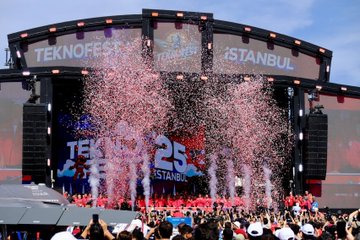 A large stage with a vibrant red backdrop displaying "TEKNOFEST 25 ISTANBUL" and logos. Pink confetti falls from above, and people in red shirts stand on stage. A crowd of attendees, some taking photos, fills the foreground. A speaker stands at a podium with a TEKNOFEST logo. Children in red jackets work on projects at a table with cardboard models and red balloons nearby.