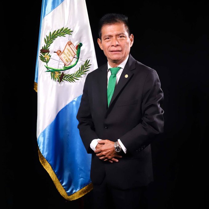 MartÃ­n NicolÃ¡s stands in a black suit and green tie, hands clasped in front. The Guatemalan flag is visible behind him, featuring blue, white, and green colors with a coat of arms.