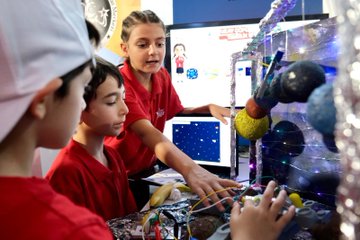 Children in red shirts working on a model with wires, lights, and a robotic arm, surrounded by a molecular structure model. Adults in red shirts observe. A screen displays a starry background. Purple stands with text and a large cube display with text are visible in the background.