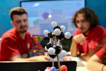 Children in red shirts working on a model with wires, lights, and a robotic arm, surrounded by a molecular structure model. Adults in red shirts observe. A screen displays a starry background. Purple stands with text and a large cube display with text are visible in the background.