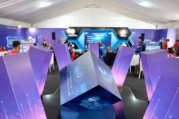 Children in red shirts working on a model with wires, lights, and a robotic arm, surrounded by a molecular structure model. Adults in red shirts observe. A screen displays a starry background. Purple stands with text and a large cube display with text are visible in the background.