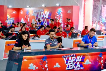 Children in red shirts working on a model with wires, lights, and a robotic arm, surrounded by a molecular structure model. Adults in red shirts observe. A screen displays a starry background. Purple stands with text and a large cube display with text are visible in the background.