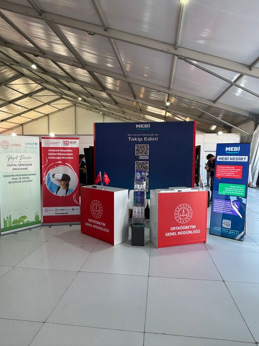 Five individuals wearing black uniforms and white hats stand in front of a blue banner with the text "MEBİ" and "Ortaöğretim Genel Müdürlüğü." The banner includes a QR code and images of digital screens. Red podiums with the logo of Genel Müdürlük are visible, along with additional banners displaying text about Yeşil Beceriler and Harezmi Eğitim Modeli. The setting is a large indoor area with a tent-like ceiling.