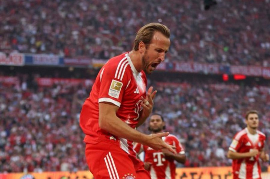 Harry Kane wearing a red Bayern Munich jersey with white stripes, celebrating on a soccer field. His mouth is open, and he appears to be shouting. Other players in similar red jerseys are visible in the background, along with a large crowd of spectators.