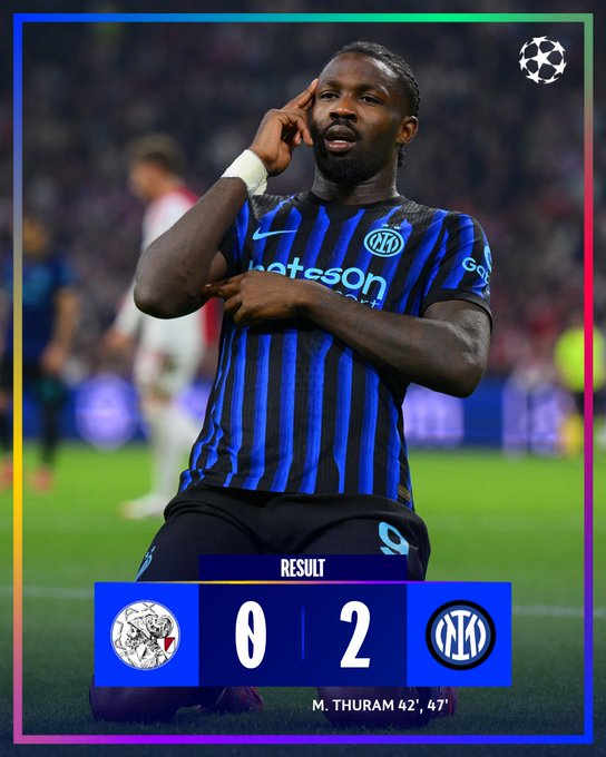 Marcus Thuram kneeling on a soccer field, wearing a blue and black striped Inter Milan jersey with "Nikon" and "Socios.com" logos. His right hand touches his head, and his left hand points forward. The background shows a stadium with blurred spectators. A score overlay displays Ajax 0, Inter 2, with Marcus Thuram\'s name and goals at 42\' and 47\'.