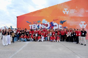 People in red jackets and shirts working on robotics projects and models at tables. A group of individuals, including children, assemble colorful robotic kits and a gingerbread house model. Displays and banners with text are visible in the background, including "Teknofest" and "Tubitak" logos.