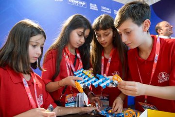 People in red jackets and shirts working on robotics projects and models at tables. A group of individuals, including children, assemble colorful robotic kits and a gingerbread house model. Displays and banners with text are visible in the background, including "Teknofest" and "Tubitak" logos.