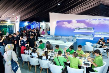 People in red jackets and shirts working on robotics projects and models at tables. A group of individuals, including children, assemble colorful robotic kits and a gingerbread house model. Displays and banners with text are visible in the background, including "Teknofest" and "Tubitak" logos.