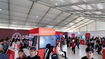 A large tent with a white canopy housing a bustling exhibition. People, including students in red and orange shirts, gather around booths with digital screens and banners. Some wear lanyards and backpacks, interacting with displays. Visible text on banners reads "T.C. Millî Eğitim Bakanlığı" and "TEKNOFEST."