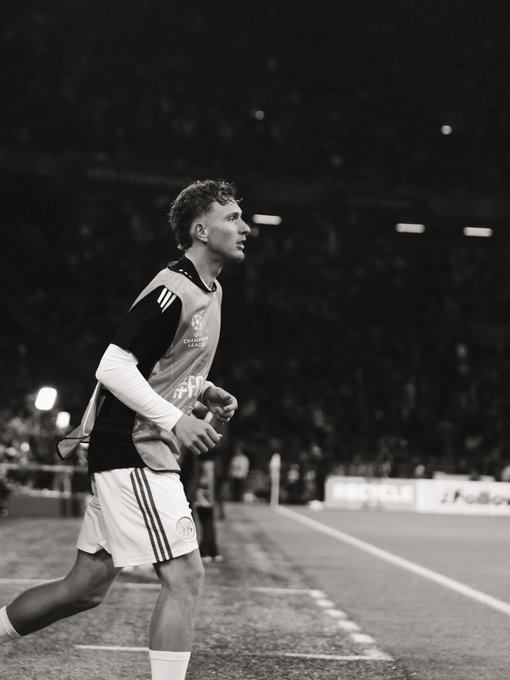 A male athlete wearing a sports jersey with long sleeves and shorts, running on a field. The jersey has dark and light patterns, and he wears socks and cleats. The background shows a stadium with blurred spectators and lights.