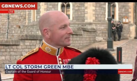 Lieutenant Colonel Storm Green MBE in a red military uniform with gold trim and a black bearskin hat with a red plume. The background shows a stone building and a street with people in uniform. Text overlay reads "LT COL STORM GREEN MBE" and "GBNEWS.COM".