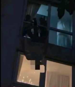 A dark, nighttime view of a multi-story building with illuminated windows. Two figures are visible outside a window on an upper floor, one appearing to hang precariously from a ledge or balcony. The scene shows a partially open window with curtains and a shadowy interior.