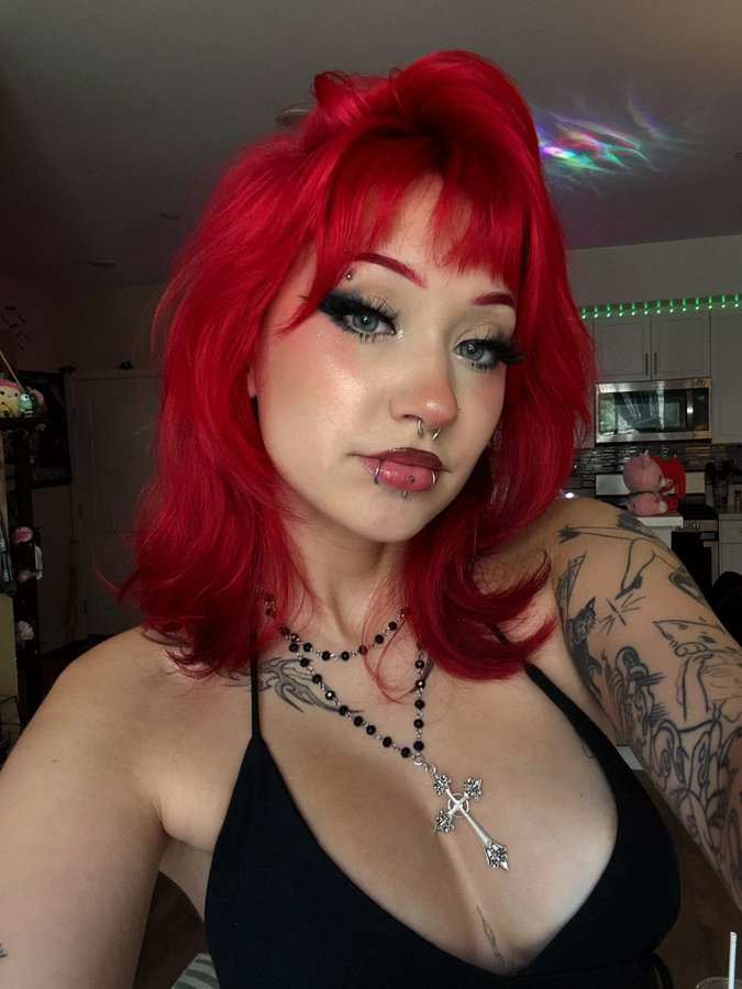 A person with vibrant red hair styled in loose waves, wearing a black sleeveless top. Visible tattoos cover the arms, featuring intricate designs. The person wears a cross necklace with black beads and has makeup including bold eyeliner and a lip piercing. The background shows a domestic setting with a kitchen and colorful lighting.