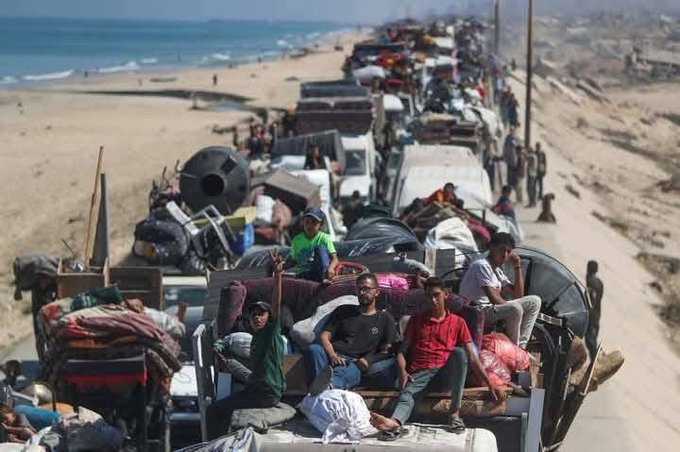 A long line of vehicles packed with people and belongings on a sandy road near a beach. People sit atop trucks and carts, surrounded by blankets, mattresses, and household items. The scene shows a crowded, dusty path with the ocean visible on one side and arid land on the other.