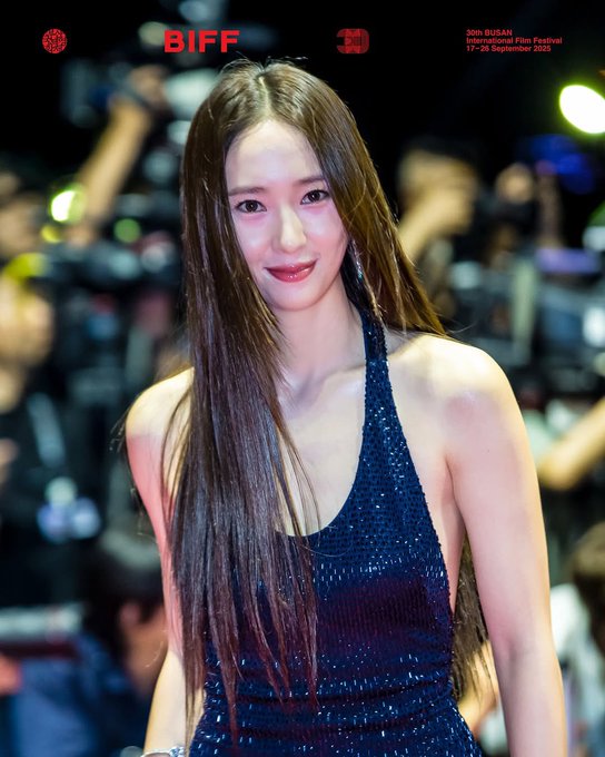 Krystal Jung wearing a sparkling dark blue halter dress with long straight hair. She stands on a red carpet with photographers and flashing lights in the background. Text overlays include "BIFF" and "30th Busan International Film Festival 5-14 September".