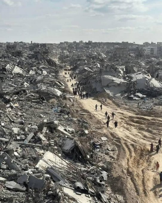 A devastated landscape with piles of rubble and debris scattered across a wide area. A dirt path runs through the center, with people walking along it. Buildings in the background are heavily damaged, with only fragments of structures remaining.