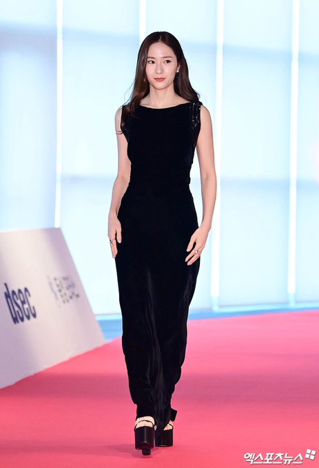 Krystal Jung walking on a red carpet in a sleeveless black velvet dress. She wears high-heeled shoes with a platform. The background includes a white banner with Korean text and photographers with cameras.