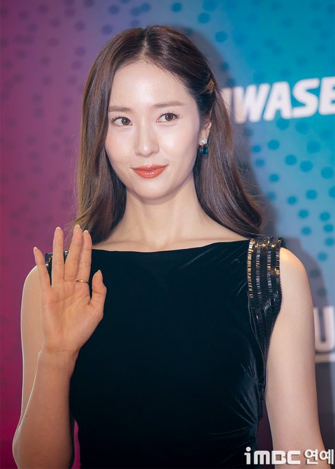 Jung Soojung stands on a stage, wearing a sleeveless black velvet dress with metallic shoulder straps. She has long brown hair and is posing with one hand raised, showing five fingers, and the other hand resting on a table with books. A colorful backdrop with the text "HWASEUN" and polka dots is visible behind her. An iMBC watermark is present in the bottom right corner.