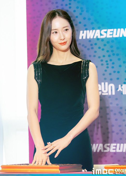Jung Soojung stands on a stage, wearing a sleeveless black velvet dress with metallic shoulder straps. She has long brown hair and is posing with one hand raised, showing five fingers, and the other hand resting on a table with books. A colorful backdrop with the text "HWASEUN" and polka dots is visible behind her. An iMBC watermark is present in the bottom right corner.