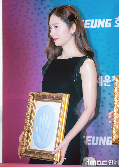 Jung Soojung stands holding a framed handprint with a blue hand design, wearing a sleeveless black dress. Her hair is styled in loose waves, and she wears earrings. The background features a colorful backdrop with text in Korean.