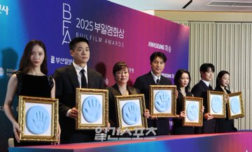 Krystal Jung stands holding a framed handprint with a blue hand imprint. She wears a black dress with sequined shoulders. Other individuals, including men and women in formal attire, also hold similar framed handprints. A backdrop displays "2025 Buil Film Awards" and "Hwasung." Watermarks from X are visible.