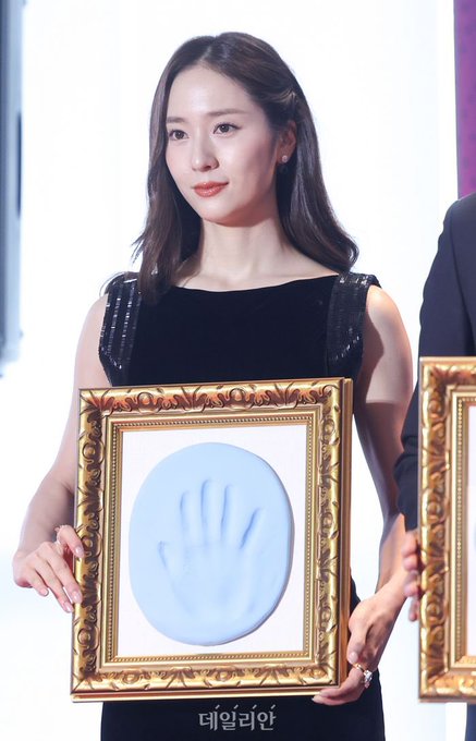 Krystal Jung stands holding a framed handprint with a blue hand imprint. She wears a black dress with sequined shoulders. Other individuals, including men and women in formal attire, also hold similar framed handprints. A backdrop displays "2025 Buil Film Awards" and "Hwasung." Watermarks from X are visible.