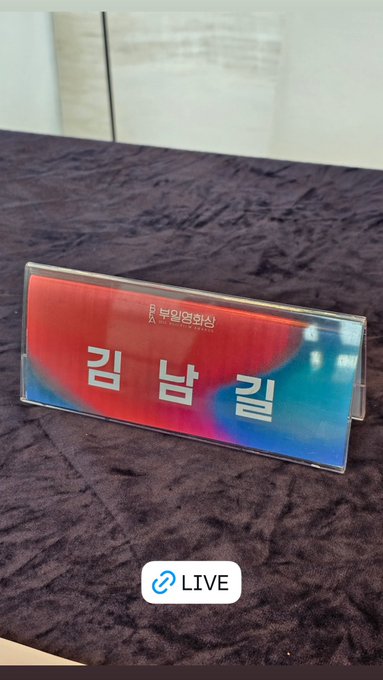 A clear acrylic nameplate on a dark table. The nameplate has a red and blue gradient background with white Korean text reading "길 남길" and smaller text above it in Korean.