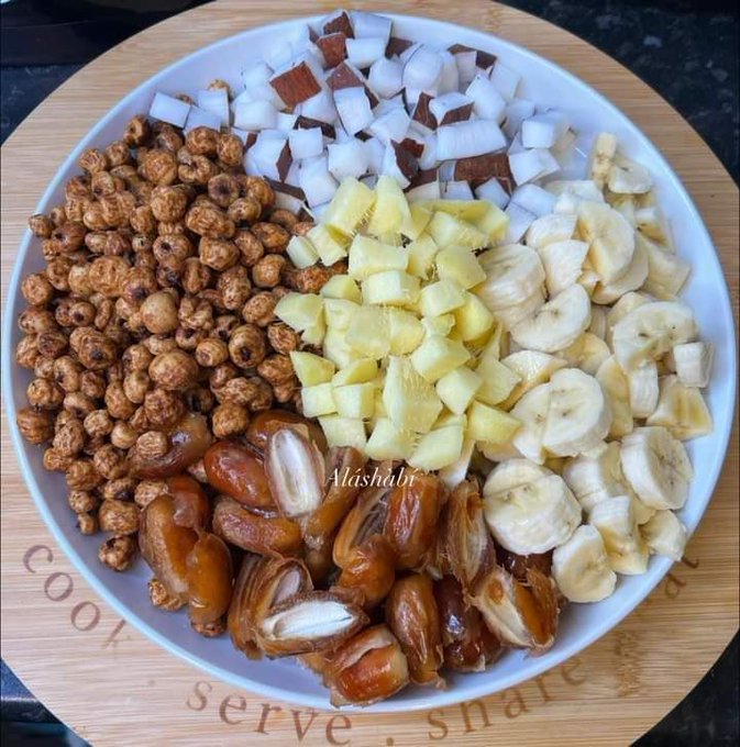 A white bowl on a wooden surface contains various ingredients. Visible are brown cereal pieces, sliced dates with pits, diced pineapple, banana slices, and coconut chunks.