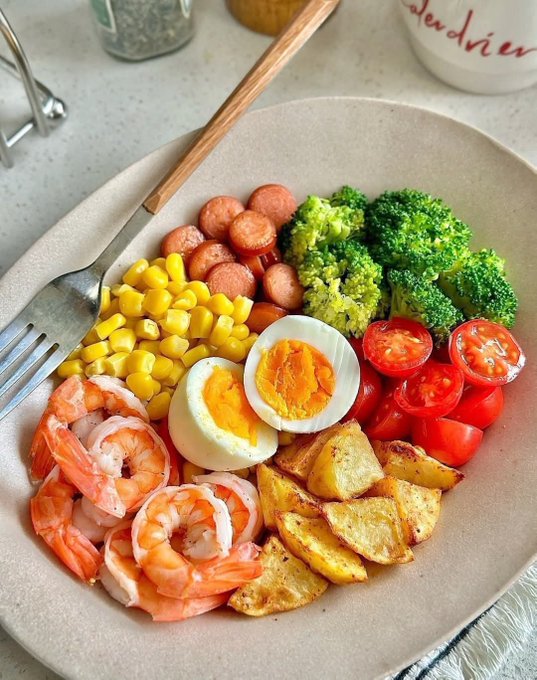 A plate of food with shrimp, boiled eggs, sliced sausages, roasted potatoes, broccoli, cherry tomatoes, and corn kernels. A wooden-handled fork and spoon are placed beside the plate on a light-colored table. A mug with the word "DRIED" and a spice container are visible in the background.