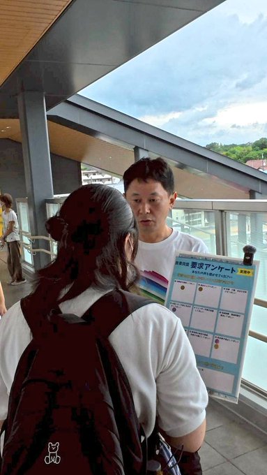Two people standing on a station platform with a modern roof and glass panels, holding a chart with text and images. One person wears a white shirt and holds the chart, while the other has a backpack. The chart includes a grid and text in Japanese, visible against a railing.