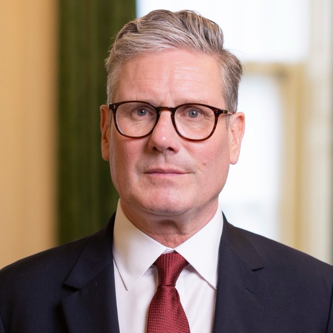 A man wearing a dark suit, white shirt, and red tie, with gray hair and glasses. A flag with horizontal green, white, and black stripes and a red triangle on the left side.