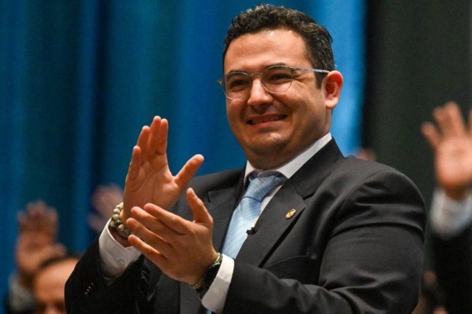 Samuel PÃ©rez clapping, wearing a dark suit, light blue tie, and glasses. His hands are raised, palms together, in a gesture of applause. Other people in the background are also clapping, with blue curtains visible behind.