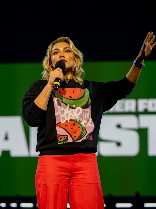 Florence Pugh stands on a stage holding a microphone in her right hand, wearing a black sweatshirt with a colorful watermelon design and the text "POWER TO THE POLLS" and red pants. Her left arm is raised, and she has a wristband on her left wrist. A green background with text is visible behind her.