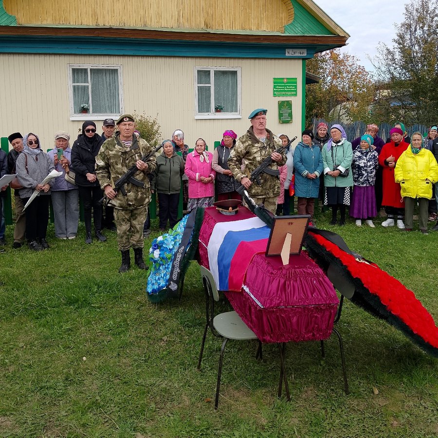 一群人，包括穿着军装、拿着步枪的人，站在一栋绿色装饰和倾斜屋顶的黄色建筑前的草地上。椅子上放着一具棺材，上面有蓝色和红色条纹，类似于俄罗斯国旗。棺材上放着一张装裱的照片，周围环绕着鲜花和花圈。人群穿着各种外套和帽子，观看着这一幕。