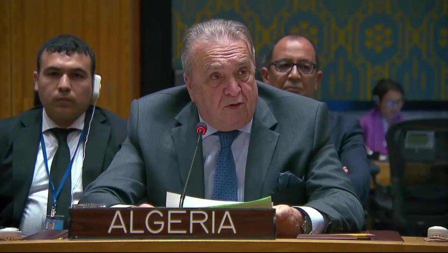 Amar Bendjama sitting at a table in a formal setting, wearing a suit and blue tie, speaking into a microphone. A nameplate reading "ALGERIA" is in front of him. Two other individuals in suits are visible in the background, seated at the table.