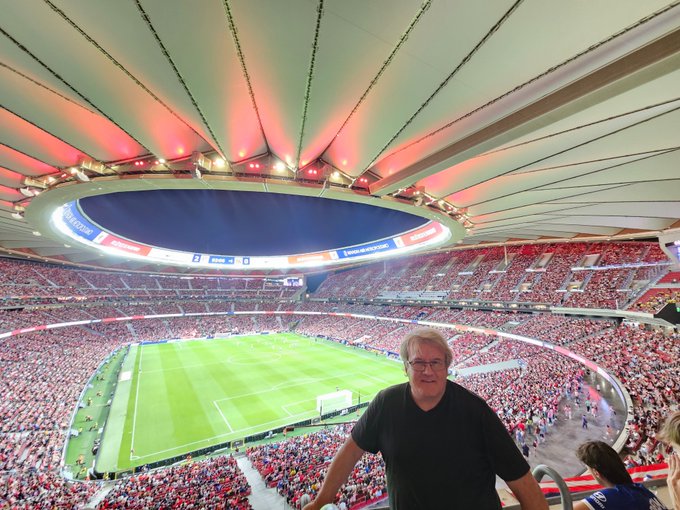 Un gran estadio lleno de espectadores, con un campo de fútbol bien iluminado en el centro. José Manuel García Suárez se encuentra en primer plano, vestido con una camiseta negra, con las gradas y el campo del Estadio Metropolitano visibles detrás de él. El estadio tiene una estructura de techo distintiva con iluminación roja.